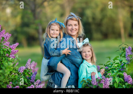 Mutter Frau mit zwei nett lächelnden Mädchen Schwestern gemeinsam schön auf einem lila Feld Bush alle tragen elegante Kleider und Mäntel Jeans Stockfoto