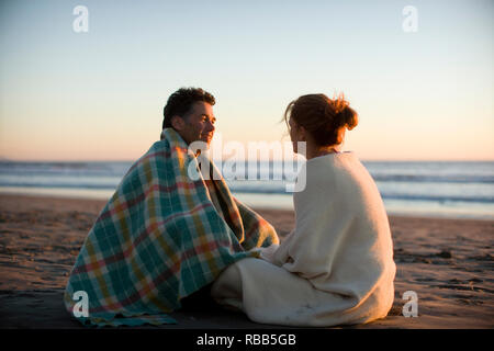 Mitte der erwachsenen Mann und seine Frau saß auf einem Strand mit Decken, um Sie bei Sonnenuntergang. Stockfoto