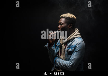 Afrikanischer Mann rauchen oder vaping E-Cig oder elektronische Zigarette Holding ein Mod mit viel Wolken isoliert auf Schwarz Stockfoto