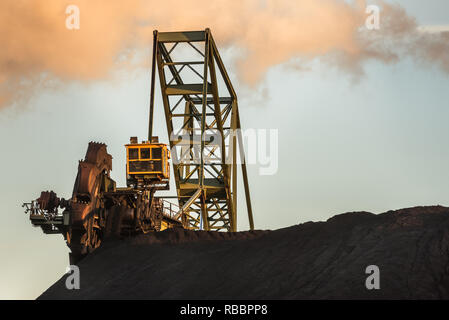 Ein schaufelradbagger ist bei der Arbeit auf einem Haufen Kohle in einem Kohlekraftwerk mit einem Rauch in den Himmel über dem Stapel. Stockfoto