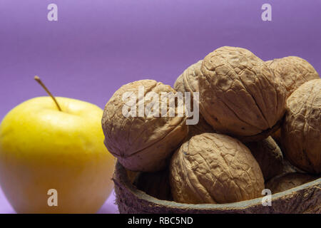 Stapel der gesamten gemeinsamen Walnüsse mit Schale in natürlichen Coconut shell Cup und gelber Apfel auf lila Hintergrund Stockfoto