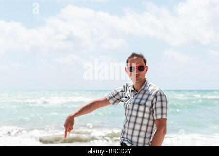 Junger Mann hipster tausendjährigen Lustig humor Gesicht am Strand während der sonnigen Tag mit roten Sonnenbrillen in Miami, Florida, mit Blick auf das Meer im Hintergrund Hände finger Po Stockfoto