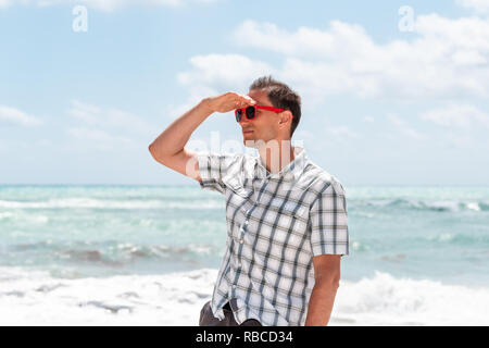 Junger Mann hipster Tausendjährigen arme Hand, die Augen von Sonne am Strand während der sonnigen Tag mit roten Sonnenbrillen in Miami, Florida, mit Blick auf das Meer im Hintergrund Stockfoto