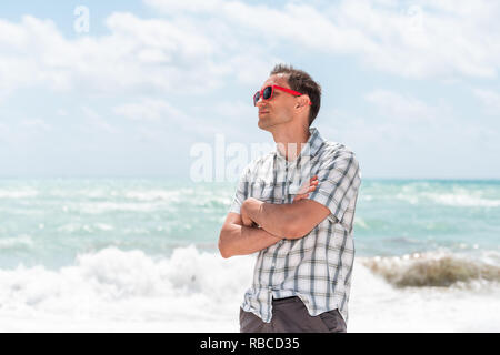 Junger Mann hipster tausendjährigen Waffen am Strand während der sonnigen Tag mit roten Sonnenbrillen in Miami, Florida, mit Blick auf das Meer im Hintergrund gekreuzt Stockfoto