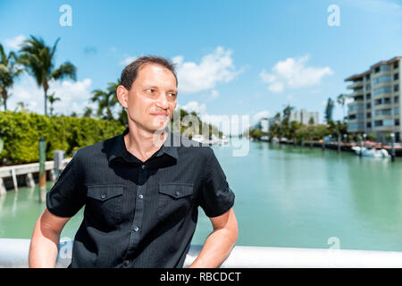 Junge Mann Gesicht mit schwarzen T-Shirt steht auf Brücke Geländer in Bal Harbour, Miami Florida lehnte sich mit Green Ocean Biscayne Bay. Stockfoto