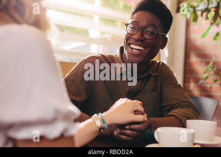 Afrikanische Kerl seiner Freundin Hand und lachte. Freundliche Paar in Liebe im Coffeeshop halten sich an den Händen sitzen. Stockfoto