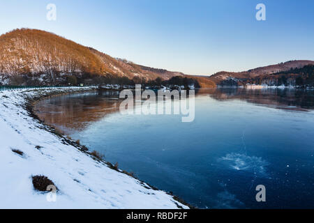 Abendlicher Blick über den zugefrorenen Obersee Rursee in der Nähe der Ortschaft Rurberg mit Schnee im Winter in der Eifel, Deutschland. Stockfoto