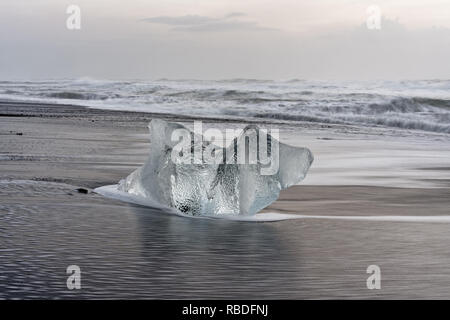 Markante Eisblock auf einem schwarzen Strand mit Surf, mit wieder Wasser Reflexion auf dem nassen Sand, Wellen im Hintergrund rolling - Ort: Island Stockfoto