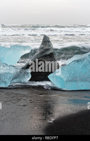 Eisblöcke in Blau und dunklen Schatten auf einem schwarzen Strand mit starker Brandung, das Eis in den nassen Sand reflektiert wird, Wellen im Hintergrund - Lage: I Stockfoto