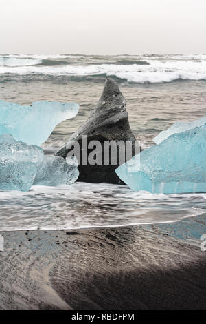 Eisblöcke in Blau und dunklen Schatten auf einem schwarzen Strand mit starker Brandung, das Eis in den nassen Sand reflektiert wird, Wellen im Hintergrund - Lage: I Stockfoto