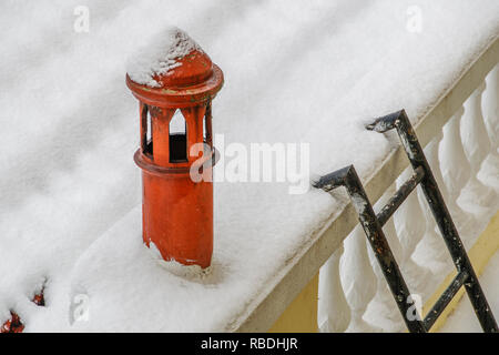 Schnee bedeckte Dach und Schornstein schließen, während ein Wintertag. Eine Metallleiter führt auf die Dachterrasse neben dem roten Schornstein Rohr. Stockfoto