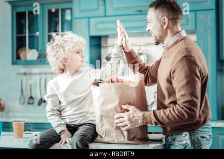 Hiermit starten. Stattliche Junge sitzt auf dem Tisch und das Berühren der Hände mit Vater, das Abendessen zu kochen Stockfoto