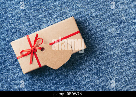 Kraftpapier Geschenkbox mit roter Schleife und Tag in Form von Herzen auf blauen Jeans. Valentinstag Karte. Stockfoto