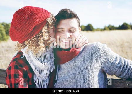 Romantische junge Paar eine schöne blonde Frau mit lockigem Haar und einen roten wolle Gap küssen zu ihrem Freund und ein stattlicher Mann outdoor Stockfoto
