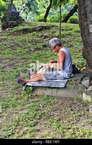 Manila, Philippines-October 24, 2016: Lokale Künstler schnitzt eine Holz- Tablet sitzen auf dem Boden unter einem Baum im Japanischen Garten des Liwasang Stockfoto