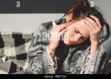 Thema Krankheit und Behandlung. jungen kaukasischen Frau sitzt home Schlafzimmer Bett im Körper Decke mit hohem Fieber, Fieber, starken Kopfschmerzen hält hinter Stockfoto