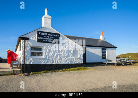 Eine Frau in Rot mit einem Hut gekleidet steht neben der Ersten und Letzten Geschenk und Erfrischung Haus in England auf einem sonnigen klaren Tag. Land's End, Cornwall. Stockfoto