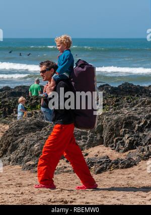 Vater zu Fuß am Strand entlang an einem sonnigen Tag und sein kleiner Sohn ein huckepack Fahrt auf Croyde Strand, Croyde, Devon, VEREINIGTES KÖNIGREICH, Stockfoto