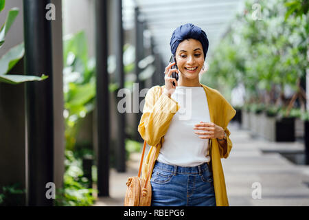 Eine junge und schöne Orientalische Frau in einem turban Hijab ist Lächeln, wie Sie auf Ihrem Smartphone die Gespräche auf der Straße während des Tages. Stockfoto