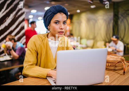 Eine junge Malay asiatische Frau, ist das Schreiben und Arbeiten oder Studieren auf Ihren Laptop in einem Cafe während des Tages. Sie trägt einen Turban Hijab. Stockfoto