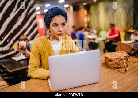 Eine junge Malay asiatische Frau, ist das Schreiben und Arbeiten oder Studieren auf Ihren Laptop in einem Cafe während des Tages. Sie trägt einen Turban Hijab. Stockfoto