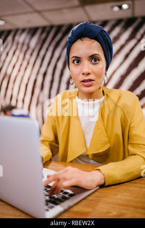 Eine junge Malay asiatische Frau, ist das Schreiben und Arbeiten oder Studieren auf Ihren Laptop in einem Cafe während des Tages. Sie trägt einen Turban Hijab. Stockfoto