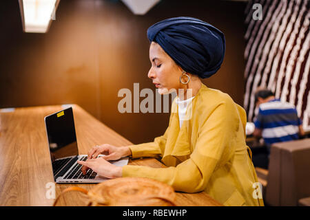 Eine junge Malay asiatische Frau, ist das Schreiben und Arbeiten oder Studieren auf Ihren Laptop in einem Cafe während des Tages. Sie trägt einen Turban Hijab. Stockfoto