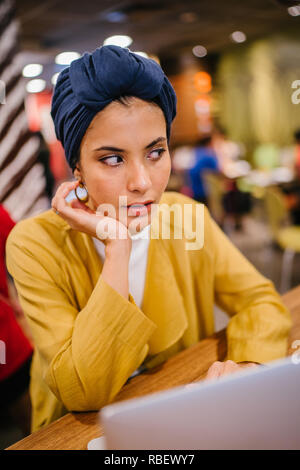 Eine junge Malay asiatische Frau, ist das Schreiben und Arbeiten oder Studieren auf Ihren Laptop in einem Cafe während des Tages. Sie trägt einen Turban Hijab. Stockfoto