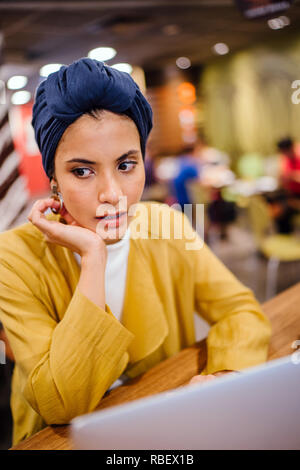 Eine junge Malay asiatische Frau, ist das Schreiben und Arbeiten oder Studieren auf Ihren Laptop in einem Cafe während des Tages. Sie trägt einen Turban Hijab. Stockfoto