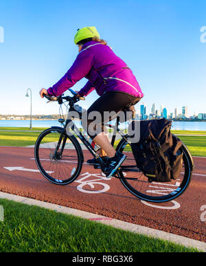 Eine Frau mittleren Alters, die mit dem Fahrrad weg-/Radweg mit einer Skyline der Stadt in der Ferne Stockfoto