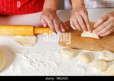 Close-up. Die ältere Frau im roten Küche Schürze rollen Sie den Teig mit einem Nudelholz, die erwachsene Tochter formt Kuchen Stockfoto