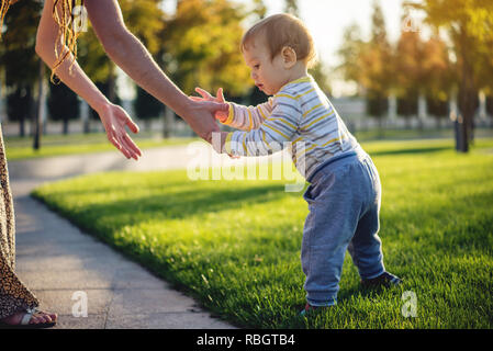 Mutter hilft niedlichen Baby zu Fuß auf einem grünen Rasen in der Natur an einem sonnigen Herbsttag. Das Konzept des ersten Kindes Schritte Stockfoto