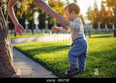 Mutter hilft niedlichen Baby zu Fuß auf einem grünen Rasen in der Natur an einem sonnigen Herbsttag. Das Konzept des ersten Kindes Schritte Stockfoto