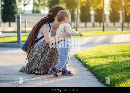 Mutter hilft niedlichen Baby zu Fuß auf einem grünen Rasen in der Natur an einem sonnigen Herbsttag. Das Konzept des ersten Kindes Schritte Stockfoto