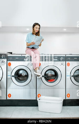 Junge Frau sitzend auf der Waschmaschine lesen Magazin beim Warten auf Wäsche waschen in den Waschsalon mit Selbstbedienung Stockfoto