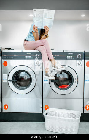 Junge Frau sitzend auf der Waschmaschine lesen Magazin beim Warten auf Wäsche waschen in den Waschsalon mit Selbstbedienung Stockfoto