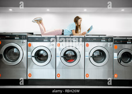 Junge Frau, die auf der Waschmaschine machiness einige Magazin lesen, während sie darauf warten, für Kleidung Wäsche in der Waschküche Stockfoto