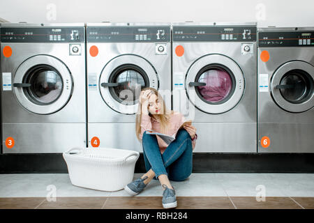 Jung und traurige Frau für die Kleidung gewaschen zu sitzen auf dem Boden im Waschsalon wartet Stockfoto