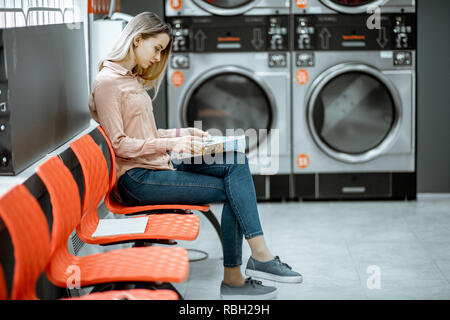 Junge Frau, die für die Kleidung gewaschen zu sitzen auf dem Stuhl bei der Waschsalon wartet Stockfoto
