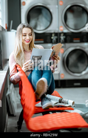 Junge Frau, die für die Kleidung gewaschen zu sitzen auf dem Stuhl bei der Waschsalon wartet Stockfoto