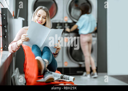 Junge Frau, die für die Kleidung gewaschen zu sitzen auf dem Stuhl bei der Waschsalon wartet Stockfoto