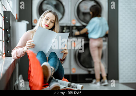 Junge Frau, die für die Kleidung gewaschen zu sitzen auf dem Stuhl bei der Waschsalon wartet Stockfoto