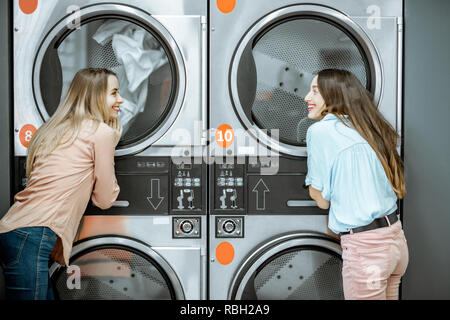 Zwei fröhliche Freundinnen stehen in unmittelbarer Nähe des Trockners Maschinen warten auf Kleidung im selbst gewartet Wäsche getrocknet werden Stockfoto