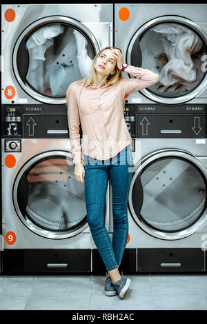 Junge Frau mit langweiligen Gefühle stehen in unmittelbarer Nähe des Trockners Maschinen für Kleidung in die Wäsche getrocknet werden, warten Stockfoto