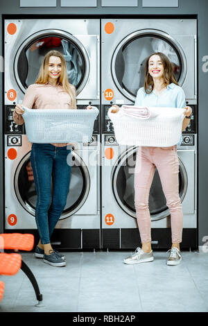 Zwei junge Frauen gemeinsam mit Körben voller Kleider in der Waschküche mit Trockner Maschinen auf dem Hintergrund Stockfoto