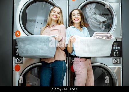 Zwei junge Frauen gemeinsam mit Körben voller Kleider in der Waschküche mit Trockner Maschinen auf dem Hintergrund Stockfoto