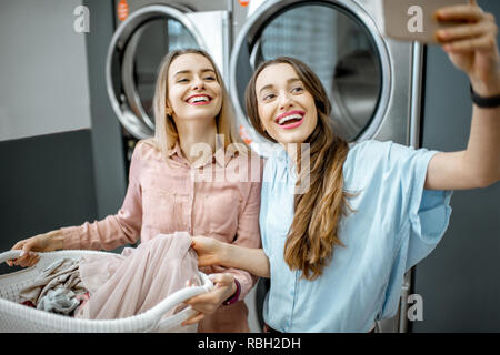 Zwei fröhliche Freundinnen machen selfie Foto mit Korb voller Kleider in der professionellen Wäscherei Stockfoto