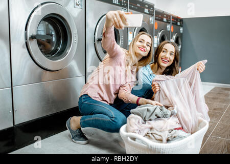 Zwei fröhliche Freundinnen machen selfie Foto mit Korb voller Kleider in der professionellen Wäscherei Stockfoto