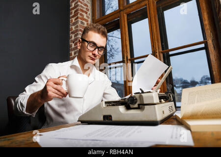 Junge Schriftsteller Brille und weißem Hemd in der Nähe der Schreibmaschine zu sitzen, Kaffee zu trinken und schreiben Neues Kapitel seines Buches in seinem Büro Stockfoto