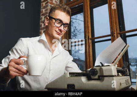 Junge Schriftsteller Brille und weißem Hemd in der Nähe der Schreibmaschine zu sitzen, Kaffee zu trinken und das Lesen neuer Kapitel seines Buches in seinem Büro Stockfoto